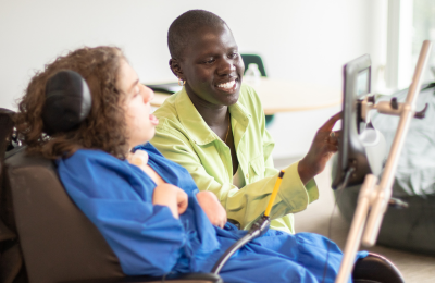 A young adult sits next to and communicates with student in blue shirt using AAC device.